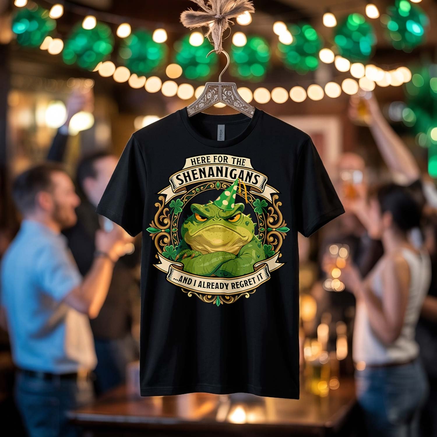 Black premium t-shirt hanging in a blurred festive pub environment, featuring a crisp vector design of a very grumpy toad in a party hat with vintage banners reading "Here For The Shenanigans... And I Already Regret It".